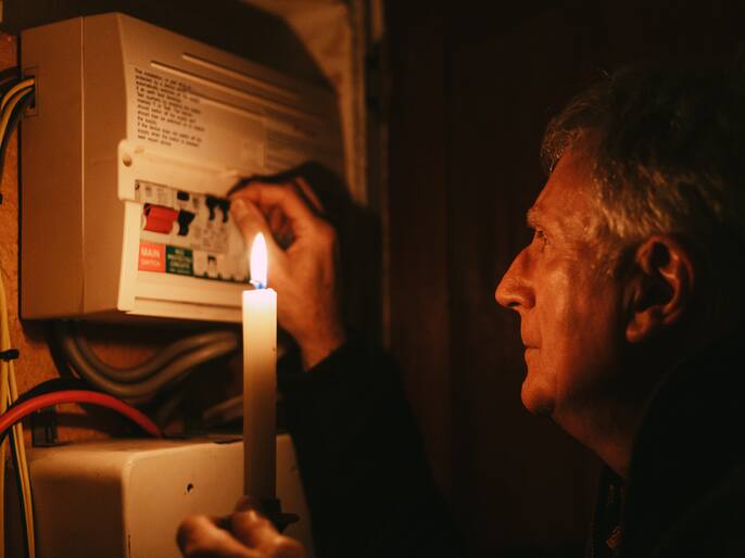A senior man investigates his fuse box at home - by the light of a candle only - in a blackout during the energy crisis.